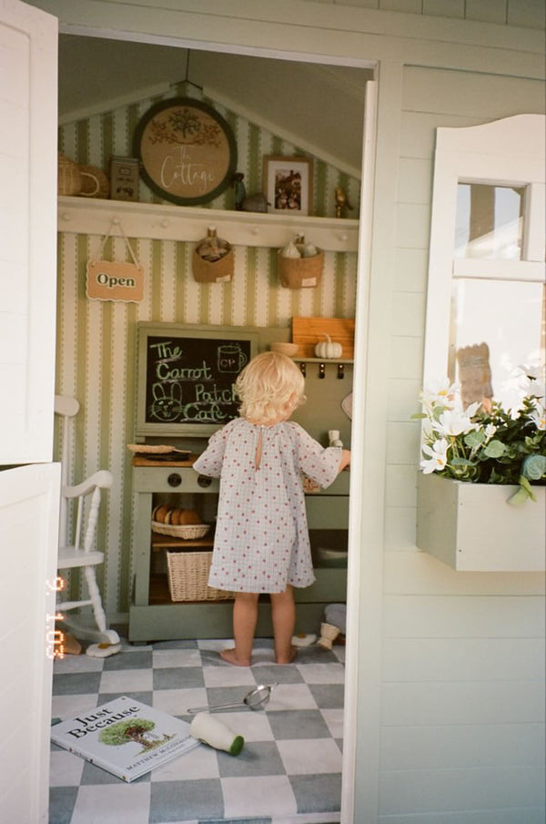 Child in a play kitchen, decorated with a chalkboard sign, patterned wallpaper, and checkered floor.